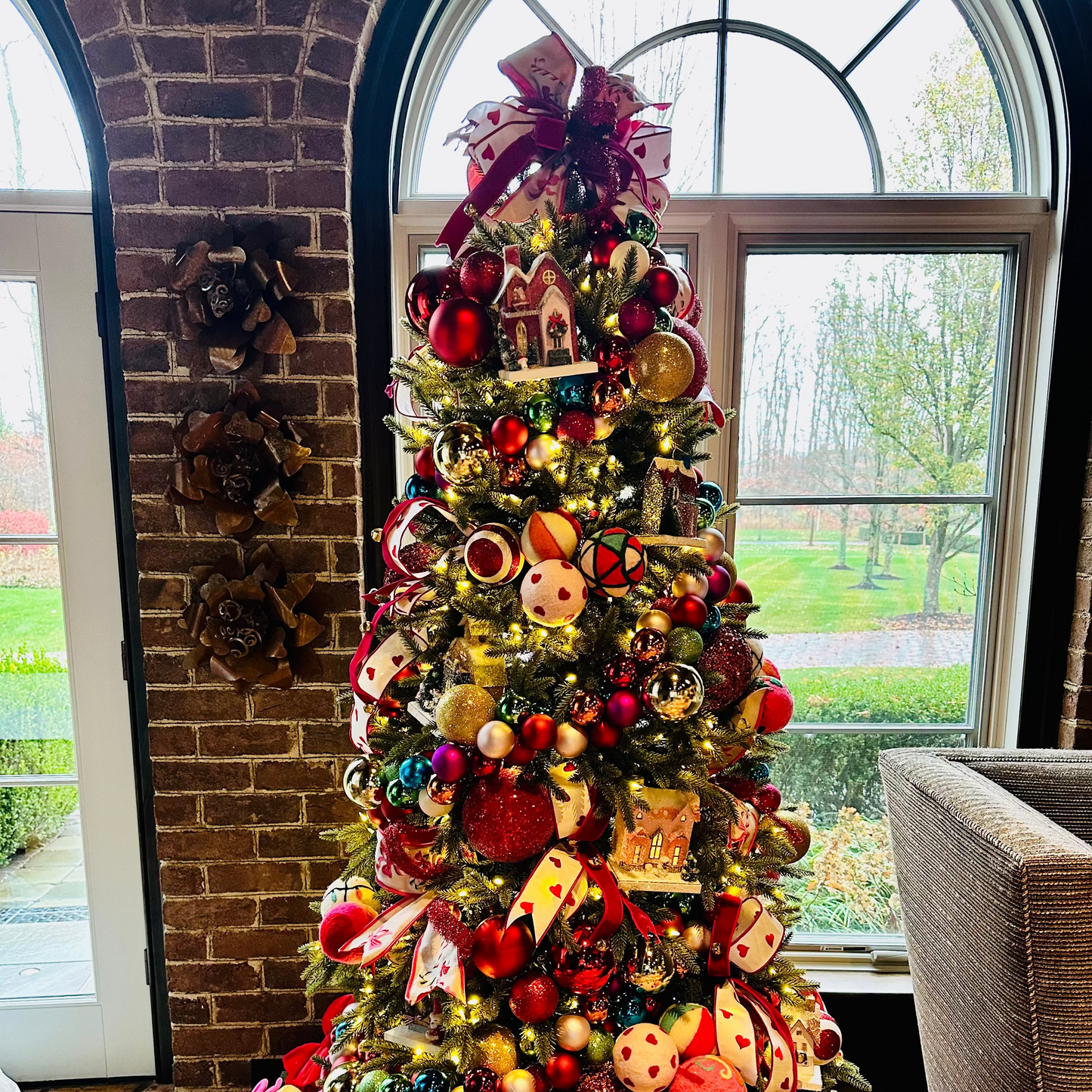 Decorated Christmas tree with colorful ornaments in a room with a brick wall and large window.