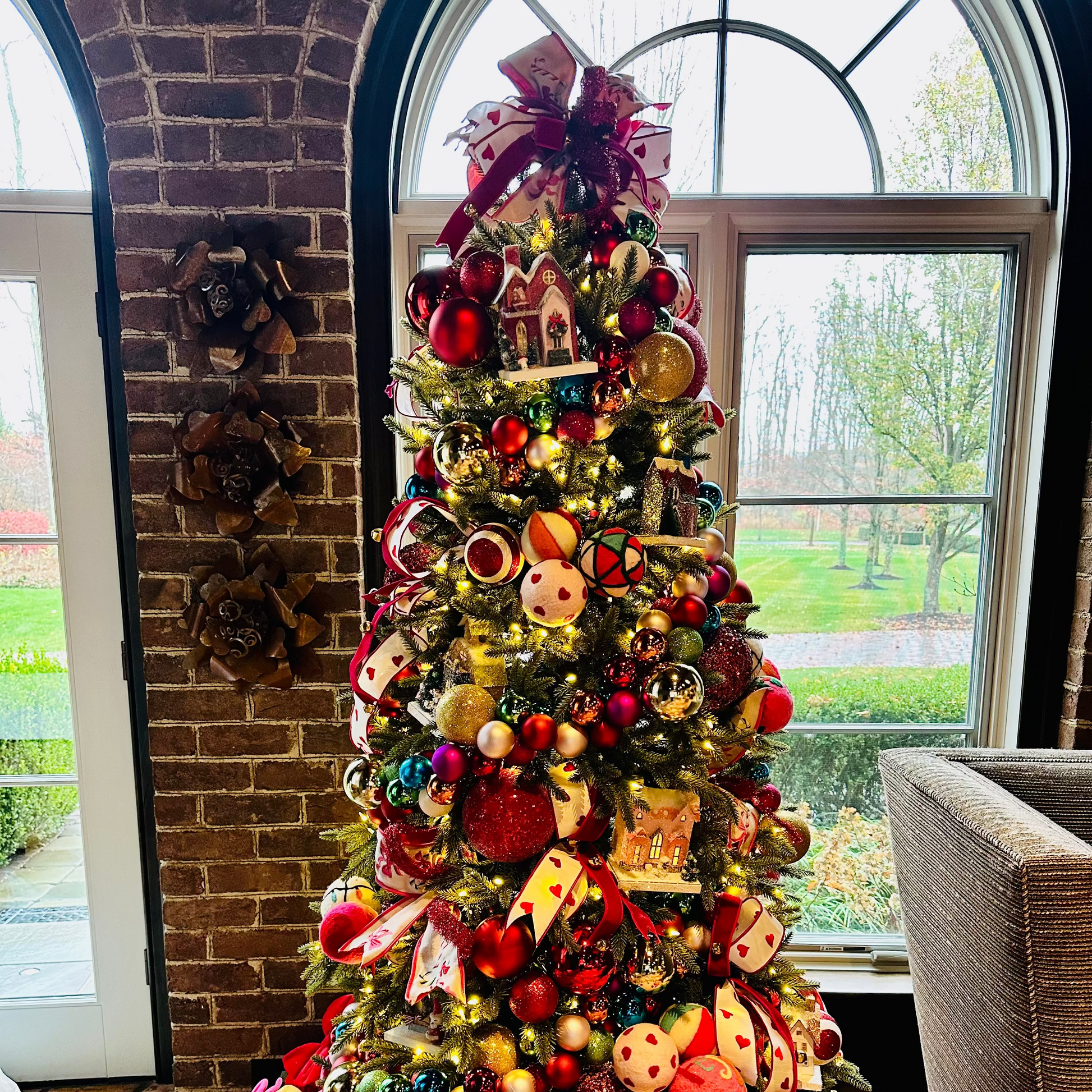 Decorated Christmas tree with colorful ornaments in a room with a brick wall and large window.